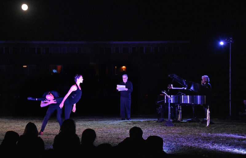 A man and woman dressed in black engage in a duet in the grass. She extends to arabesque and he wraps his arms and chest around her extended leg.
